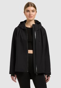 Giovane donna che indossa una giacca nera con cappuccio e cerniera sopra un top corto nero e pantaloni, in piedi davanti a uno sfondo chiaro e uniforme.