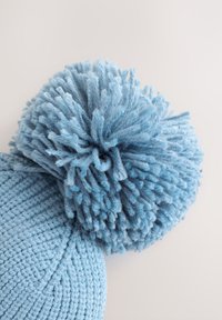Svetlomodra pletena zimska kapa z velikim, puhastim cofom iz preje na vrhu, na enostavni svetli podlagi.