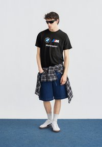 Νέος άνδρας με μαύρο μπλουζάκι BMW Motorsport, σκούρα γυαλιά ηλίου, καρό πουκάμισο δεμένο στη μέση, τζιν σορτς, λευκές κάλτσες και λευκά αθλητικά παπούτσια.