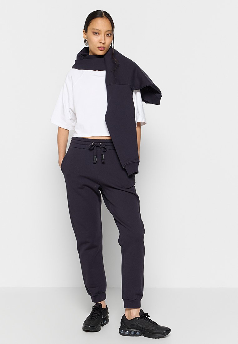 Skrócony biały t-shirt i ciemnogranatowe dresy ze sznurkiem. Model nosi ciemne sneakersy; granatowy sweatshirt zarzucony na ramionach.