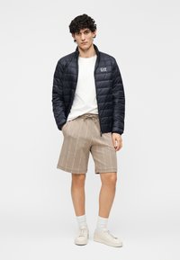 Jachetă neagră matlasată cu guler ridicat, tricou alb, pantaloni scurți bej cu un model subtil cu logo și adidași de culoare deschisă.