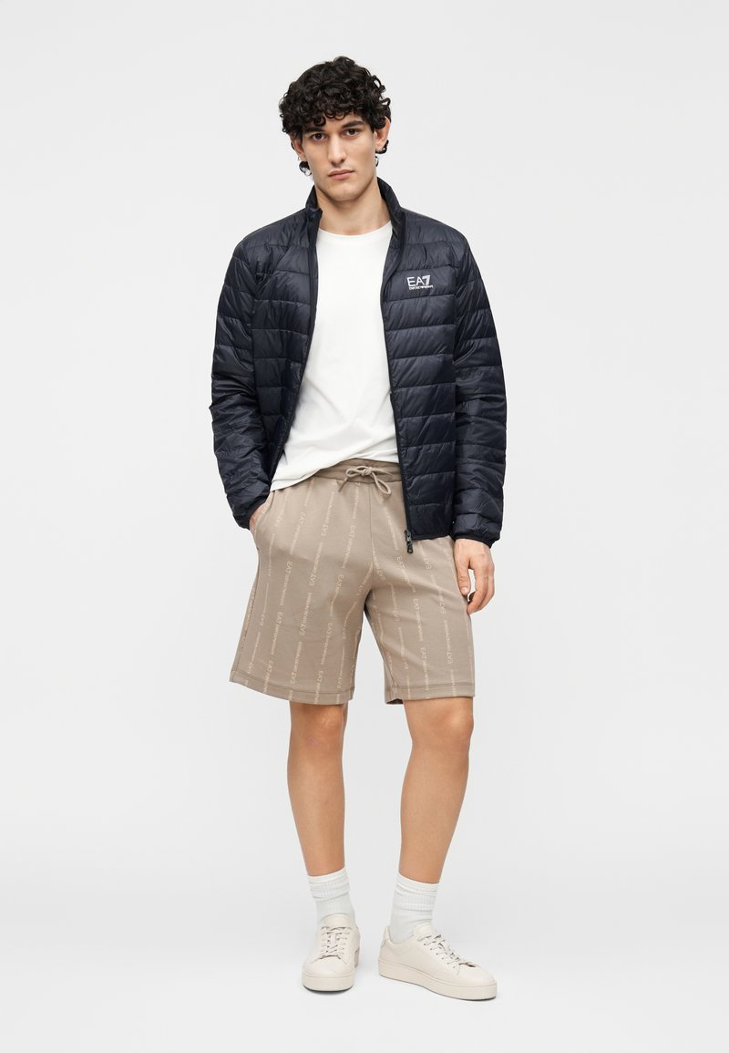 Jachetă neagră matlasată cu guler ridicat, tricou alb, pantaloni scurți bej cu un model subtil cu logo și adidași de culoare deschisă.