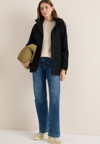 Svart vattentät jacka med dragkedja framtill, åtsittande huva och två framfickor. Bärs över en beige stickad tröja och ihop med blå vida jeans.