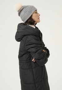 Manteau noir en duvet avec une capuche, présentant un design matelassé et une fermeture éclair. Associé à un bonnet en tricot gris clair avec un pompon en fourrure sur le dessus.