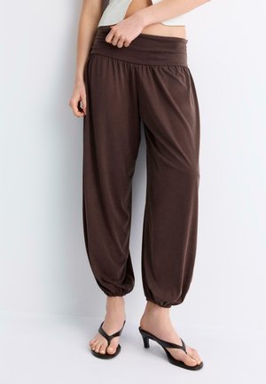 Osoba nosząca luźne, brązowe spodnie z elastycznymi mankietami, w połączeniu z czarnymi sandałami na obcasie, stojąca przy gładkiej jasnej ścianie.