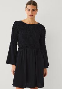 Zwarte jurk met een gestructureerde, smock-buste en uitlopende mouwen. Het heeft een ronde halslijn en een vloeiende rok, gemaakt van lichte stof.