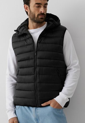 Mężczyzna w czarnej pikowanej kamizelce z kapturem na białej koszuli z długim rękawem, z ręką w kieszeni jasnoniebieskich dżinsów, patrzy w dół na bok.