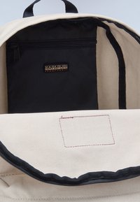 Beige plátěný batoh se vnitřkem černou zipovou kapsou a černou nálepkou. Horní část má tmavé lemování a nezašitou obdélníkovou plochu.