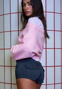 Pulover roz supradimensionat cu guler ribbit, asortat cu pantaloni scurți din denim închis, având buzunare pe spate. Modelul stă în fața unui perete cu faianță.