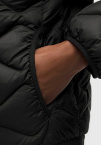 Mano de piel oscura parcialmente insertada en el bolsillo lateral de una chaqueta negra acolchada con detalles de puños elásticos.