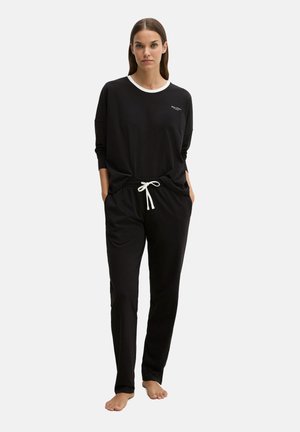 Vrouw draagt een zwart longsleeve met witte kraag en bijpassende zwarte broek met witte aantrekkoord, staat blootsvoets tegen een effen achtergrond.