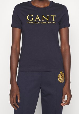 Personne portant un T-shirt GANT bleu marine avec un logo jaune et un pantalon bleu marine assorti avec un emblème jaune sur la cuisse supérieure.