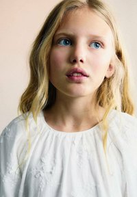 Vit blus med blommig brodyr, lång vågig blont hår, en neutral bakgrund och blå ögon. Mjukt ljus lyser upp ansiktsdragen.