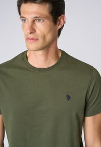 T-shirt in cotone verde oliva con colletto tondo, con un piccolo logo blu navy sul lato sinistro del petto. Tessuto liscio, aderente e maniche corte.