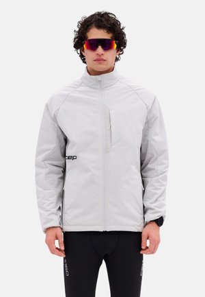 Uomo con giacca grigio chiaro con zip, pantaloni da ciclismo neri, occhiali sportivi riflettenti e uno smartwatch, in piedi davanti a uno sfondo neutro.