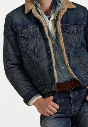 Homme portant une veste en jean bleu avec une doublure beige en shearling, une chemise bleue, une couche intérieure blanche, une ceinture marron et un jean bleu, main dans la poche.