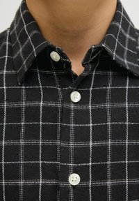 Melnā flaneļa krekls ar baltu režģa rakstu, ar klasisku apkakli, divām baltām pogām un maigu, strukturētu audumu.