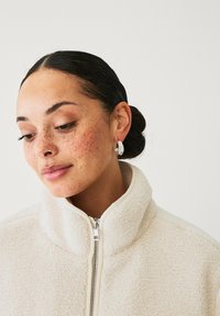 Hvid fleece zip-up jakke med høj krave. Sølv teksturerede hoops øreringe med en lille accentsten. fregner er synlige på huden.