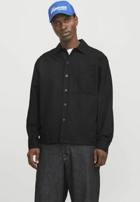 Chemise noire à boutons avec une poche, faite de tissu léger. Coupe légèrement ample, associée à un pantalon en denim foncé. Casquette bleue avec logo.