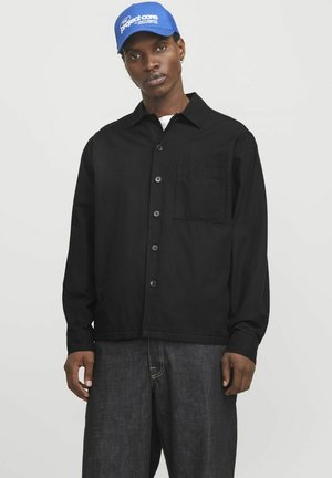 Schwarzes Hemd mit Knopfleiste und Tasche, aus leichtem Stoff. Leicht lockere Passform, kombiniert mit dunklen Jeans. Blauer Hut mit Logo.