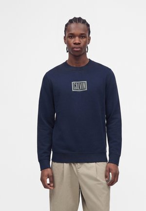 Giovane uomo con i capelli intrecciati che indossa una felpa blu navy con il logo "CALVIN" e pantaloni beige, in piedi davanti a uno sfondo bianco uniforme.