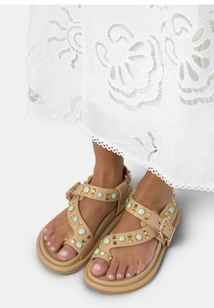 Piedi con sandali beige decorati con borchie turchesi e dorate, abbinati a una gonna bianca con ricamo a fiori traforato.