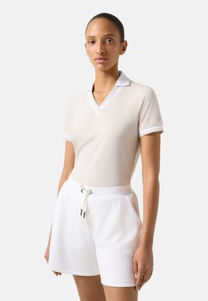 Frau trägt ein beigefarbenes kurzärmeliges V-Ausschnitt-Shirt mit weißem Kragen und weiße Shorts mit Kordelzug, steht vor einem schlichten hellen Hintergrund.
