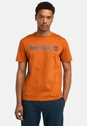 Orange cotton t-shirt featuring a blue Timberland logo and tree graphic. Round neck and short sleeves, paired with dark pants.