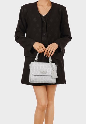 Donna con blazer e gonna neri che tiene davanti a sé una piccola borsa bianca con monogramma decorato e manici superiori.