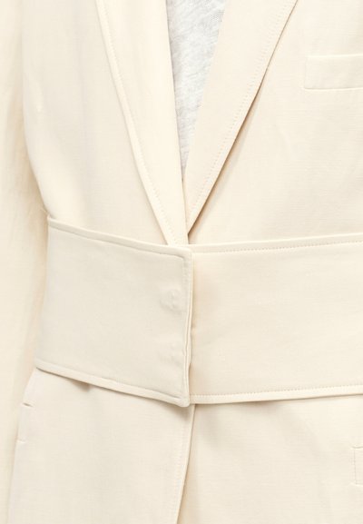 Κοντινό πλάνο μπεζ σακακιού με φαρδιά ζώνη από ίδιο ύφασμα και πέτο με σχισμή πάνω από ανοιχτό γκρι εσώρουχο.