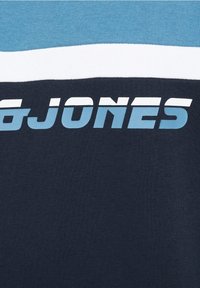 Lyhythihainen tummansininen T-paita, jossa on vaaleansiniset ja valkoiset vaHorizontal Stripe -raitaa, ja rintaosassa rohkea "JONES"-teksti vaaleansinisellä.