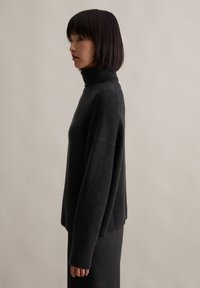 Schwarzer Rollkragenpullover aus weichem, strukturiertem Stoff mit lockerer Passform. Mit langen Ärmeln und gerippten Bündchen, kombiniert mit einem schlichten schwarzen Rock.