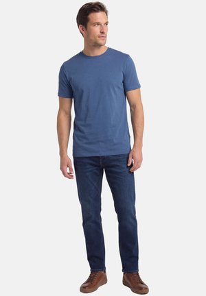 Giovane uomo con indosso una maglietta blu semplice, jeans blu scuro e sneaker di pelle marrone, in piedi e che guarda leggermente a sinistra.