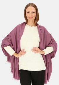 Fioletowy szal z miękkiego materiału z frędzlami, zarzucony na kremowy sweter. Modelka pozuje z rękami na biodrach na neutralnym tle.