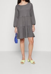 Kvinna i en lös grå lagd klänning, som håller en liten blå handväska och bär ljusgröna öppna klackskor med blockklack.