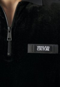 Haut en velours noir avec une fermeture éclair à l'avant et une étiquette "Versace Jeans Couture" sur la poitrine.
