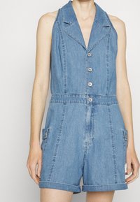 Denim overal s límečkem, pěti knoflíky, dvěma bočními kapsami, ohrnutými rukávy a světle modrou barvou s hladkou texturou.