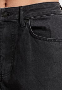 Blugi din denim negru, cu talie înaltă, buzunar frontal, închidere cu nasturi și cusături rezistente. Țesătura are o textură fină, cu un luciu ușor.