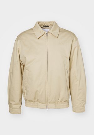 Beige zip-jakke med krage, elastiske mansjetter og midje. Har sidelommer og en subtil logo på brystet. Glatt tekstur.