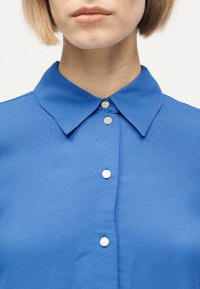 Κοντινό πλάνο ενός ατόμου που φοράει μπλε πουκάμισο με κουμπιά, με μυτερό γιακά και λευκά κουμπιά, κόβοντας από τη μύτη και κάτω.