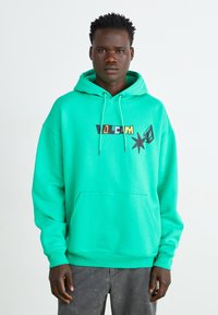 Grön hoodie med en framficka och snodd i huvan. Har grafisk text och former i svart och gult på bröstet. Tillverkad av en bomullsblandning.