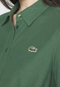Chemise verte à col avec un petit logo crocodile vert brodé sur la poitrine gauche, boutonnée près du cou.
