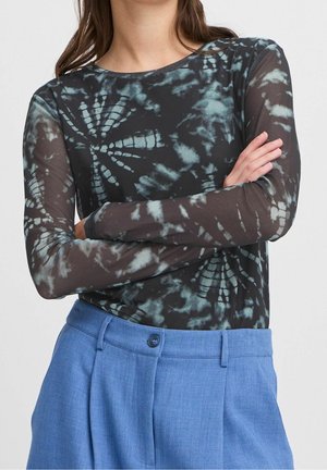 Personne portant un haut tie-dye noir et bleu à manches longues, les bras croisés, associé à un pantalon taille haute bleu clair avec un bouton.