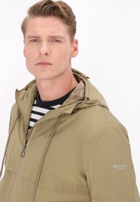 Jachetă ușoară de culoare verde măsliniu, cu glugă, fermoar frontal și șnururi de ajustare. Prezintă o textură fină și un accent de marcă pe mânecă.