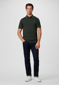 Man in een donkergroen polo shirt met gestreepte kraag en manchetten, donkere spijkerbroek en witte sneakers, staand met één hand in zijn zak.