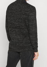 Maglione nero a trama con motivo mélange, maniche lunghe e collo alto, abbinato a jeans scuri. Materiale in maglia morbida.