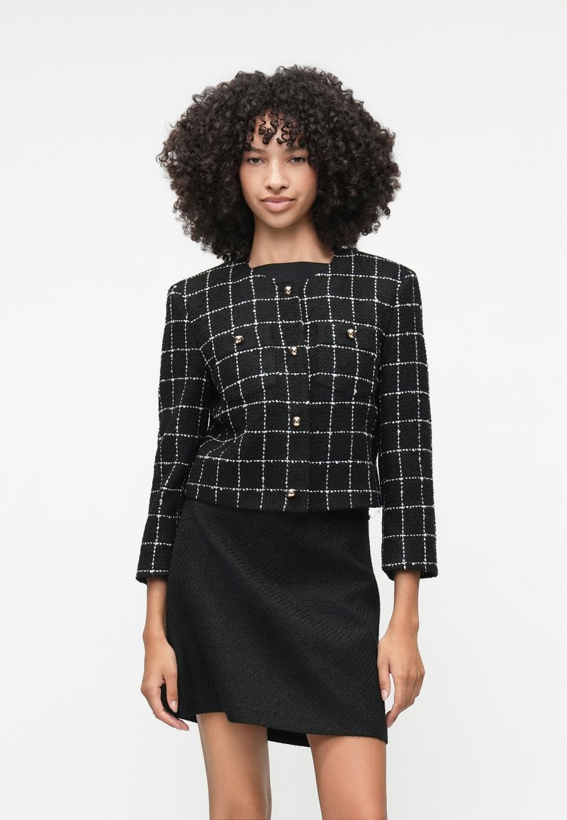 Sort cropped jakke med hvidt rutenetmønster, guldknapper og 3/4 ærmer, parret med en sort tekstureret nederdel.