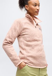 Γυναίκα που φοράει ροζ φωτεινό φλίς πουλόβερ με φερμουάρ στο ένα τέταρτο, με τα χέρια στις τσέπες, κοιτάζει προς τα δεξιά μπροστά σε απλό φωτεινό φόντο.