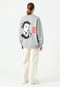 Felpa grigia con la grafica di una donna sulla schiena, con la didascalia "OOPS NON MI IMPORTA", abbinata a pantaloni cargo color crema e sneaker bianche chunky.