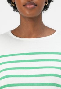 Pull blanc avec des rayures horizontales vertes, doté d'un col rond et d'une texture douce. De grandes boucles d'oreilles en or circulaires sont visibles.